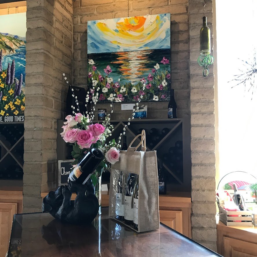 A wine display with a floral arrangement, painting, and decorative bottle holder on a table in a cozy wine shop or restaurant. The painting depicts a sunset over water with flowers in the foreground.