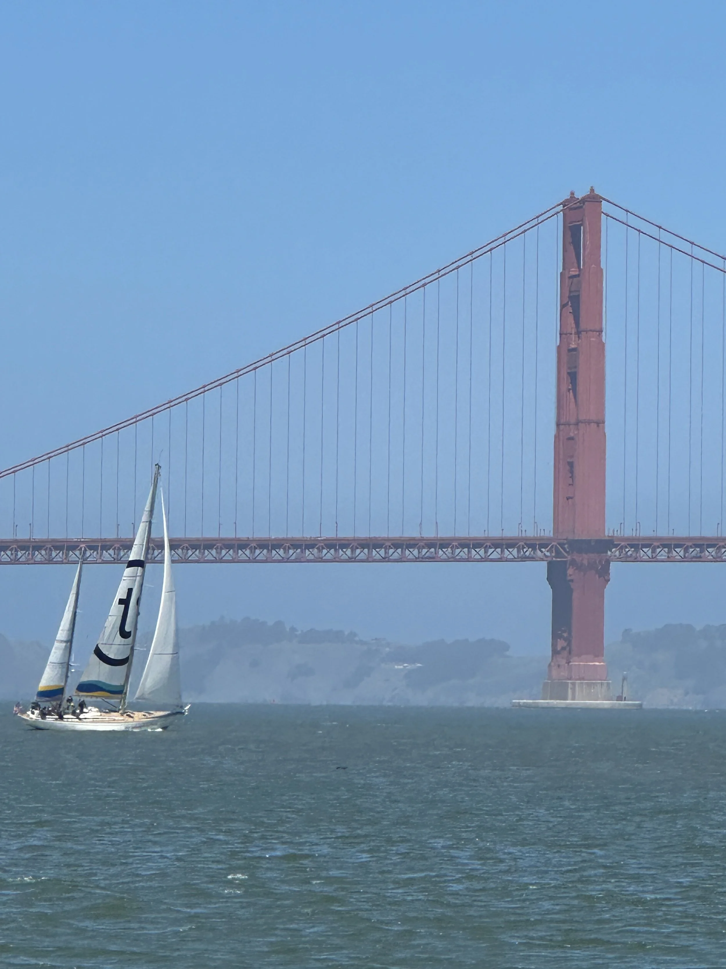 A sailboat with white sails on the water under the Golden Gate Bridge in San Francisco with a clear blue sky.