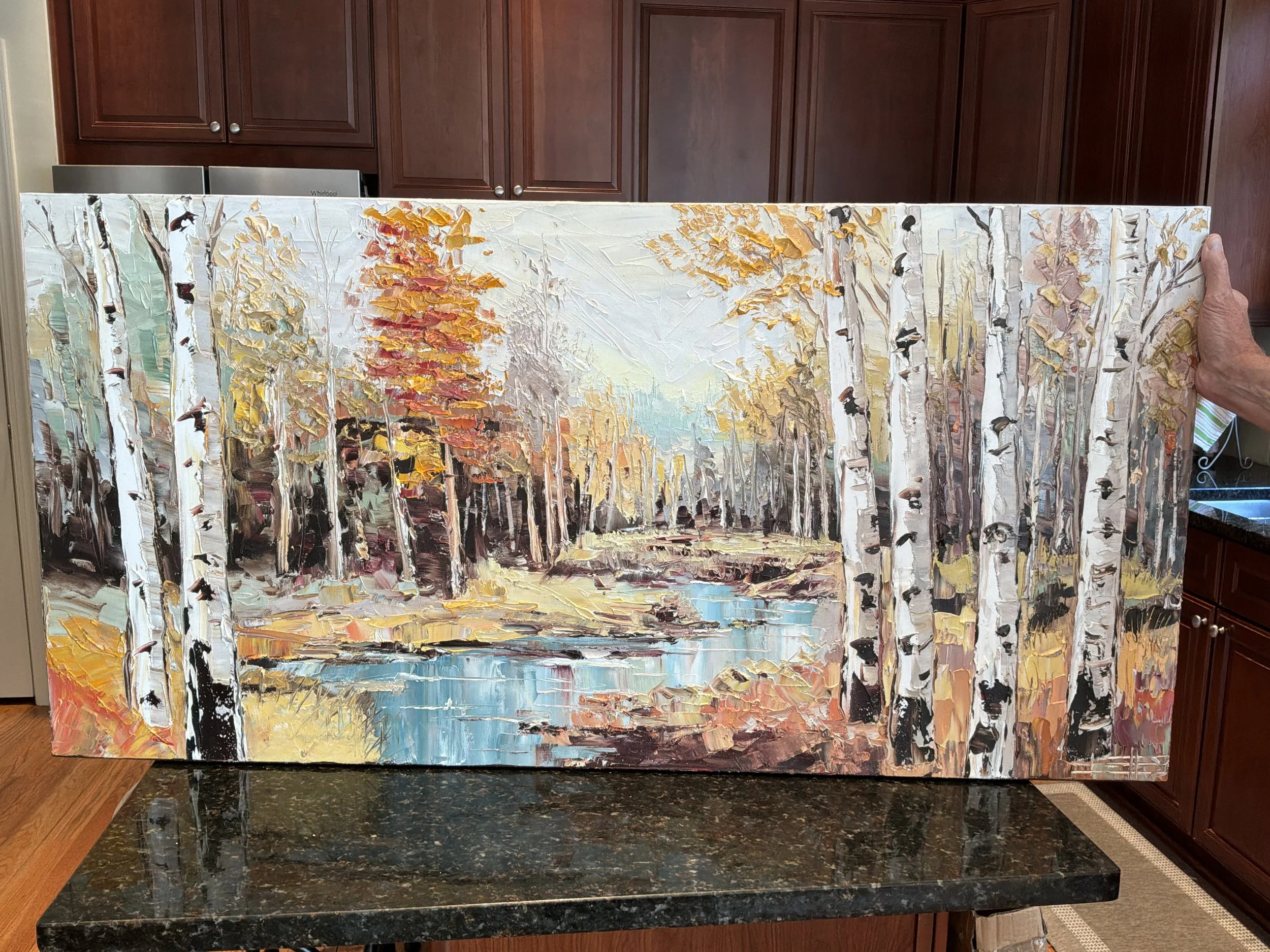 A person holding a landscape painting of a forest scene with trees and a small pond, using thick impasto brushstrokes. The background shows upper kitchen cabinets made of dark wood.