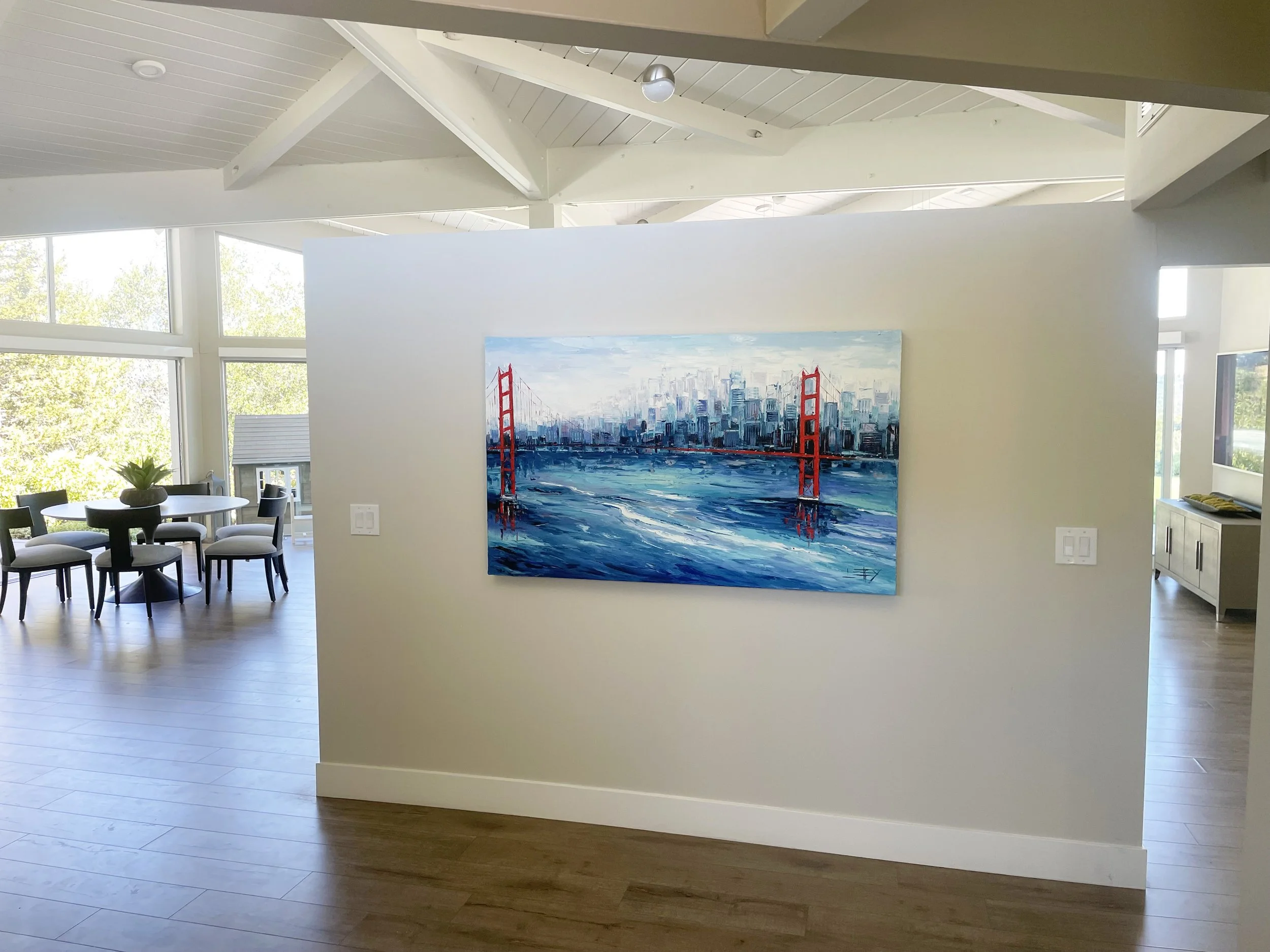 Interior of a modern house with high vaulted ceilings, large windows, a dining table with six chairs, and a large painting of the San Francisco skyline and Golden Gate Bridge on a white wall.