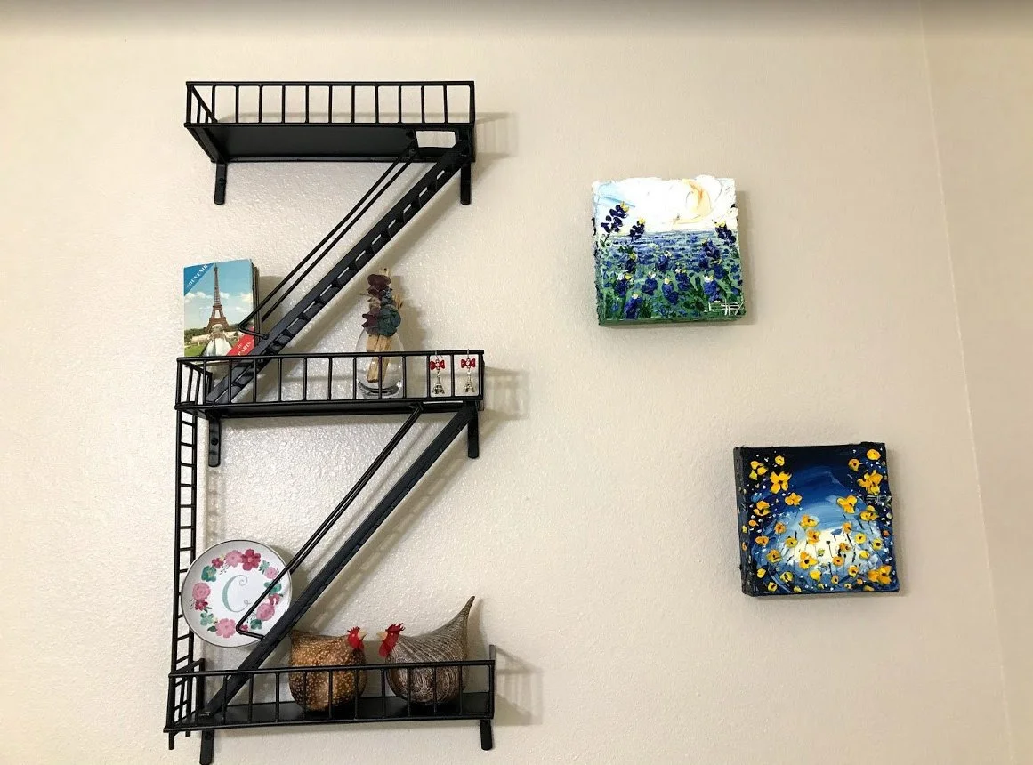 Decorative black metal wall shelf with four levels on a white wall, holding various figurines, a plate, and a book. Two small paintings of floral and landscape scenes are hung on the wall next to the shelf.