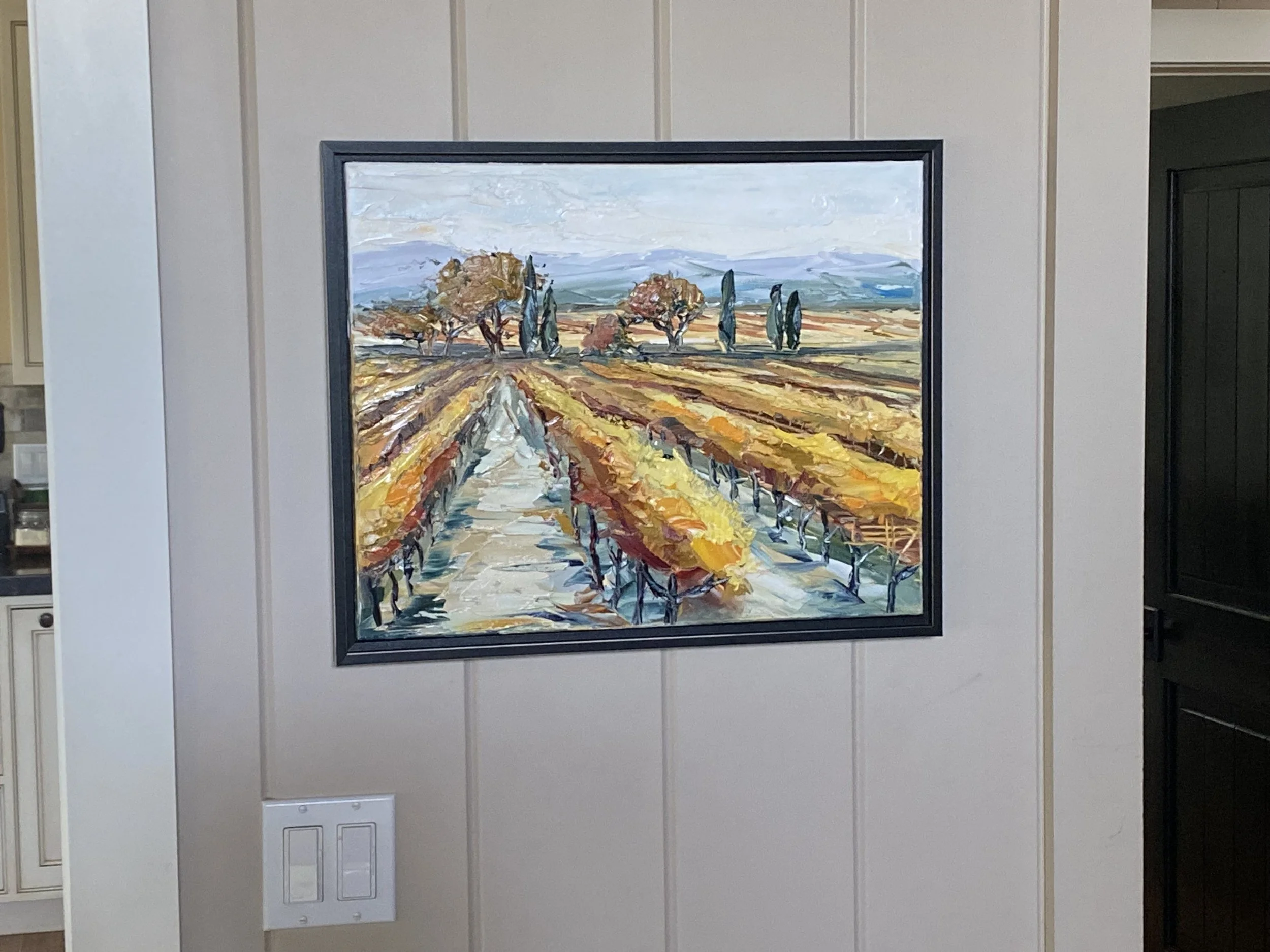 Original painting of a vineyard landscape with trees, fields, and mountains in the background, seen hanging on a wall in a room.