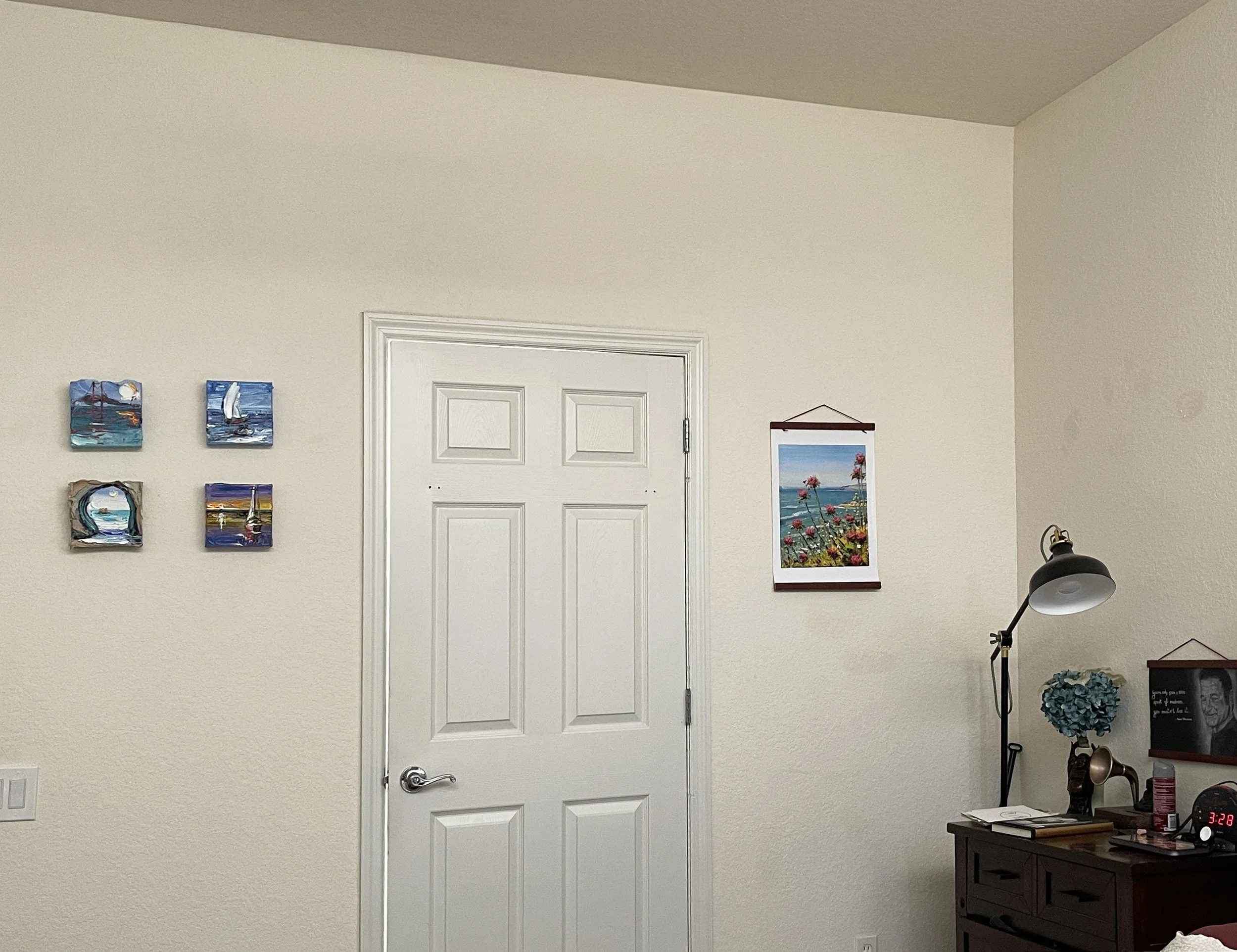 Interior view of a beige wall with a white door in the center, decorated with several small paintings and a larger framed seaside photo, next to a lamp and small table.
