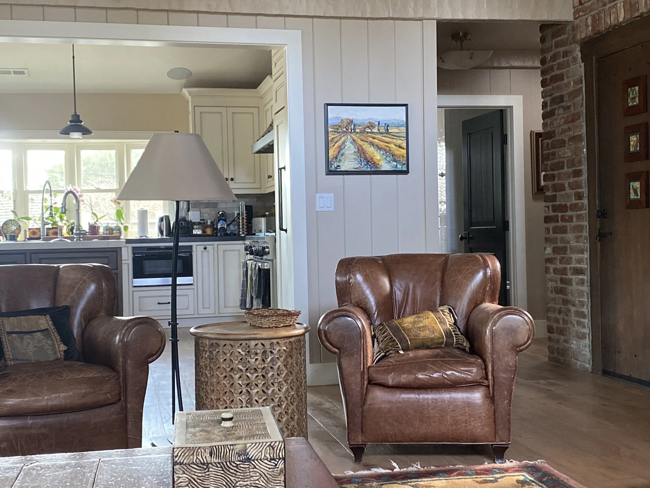 Living room with leather armchairs, a patterned side table, and a lamp. Kitchen area with white cabinets, window, and counter in the background. Wall art, brickwall, and doorway also visible.