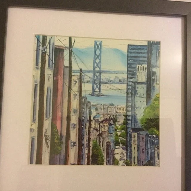 A framed watercolor painting of a city street with buildings on both sides, utility poles, and a bridge over water in the background.