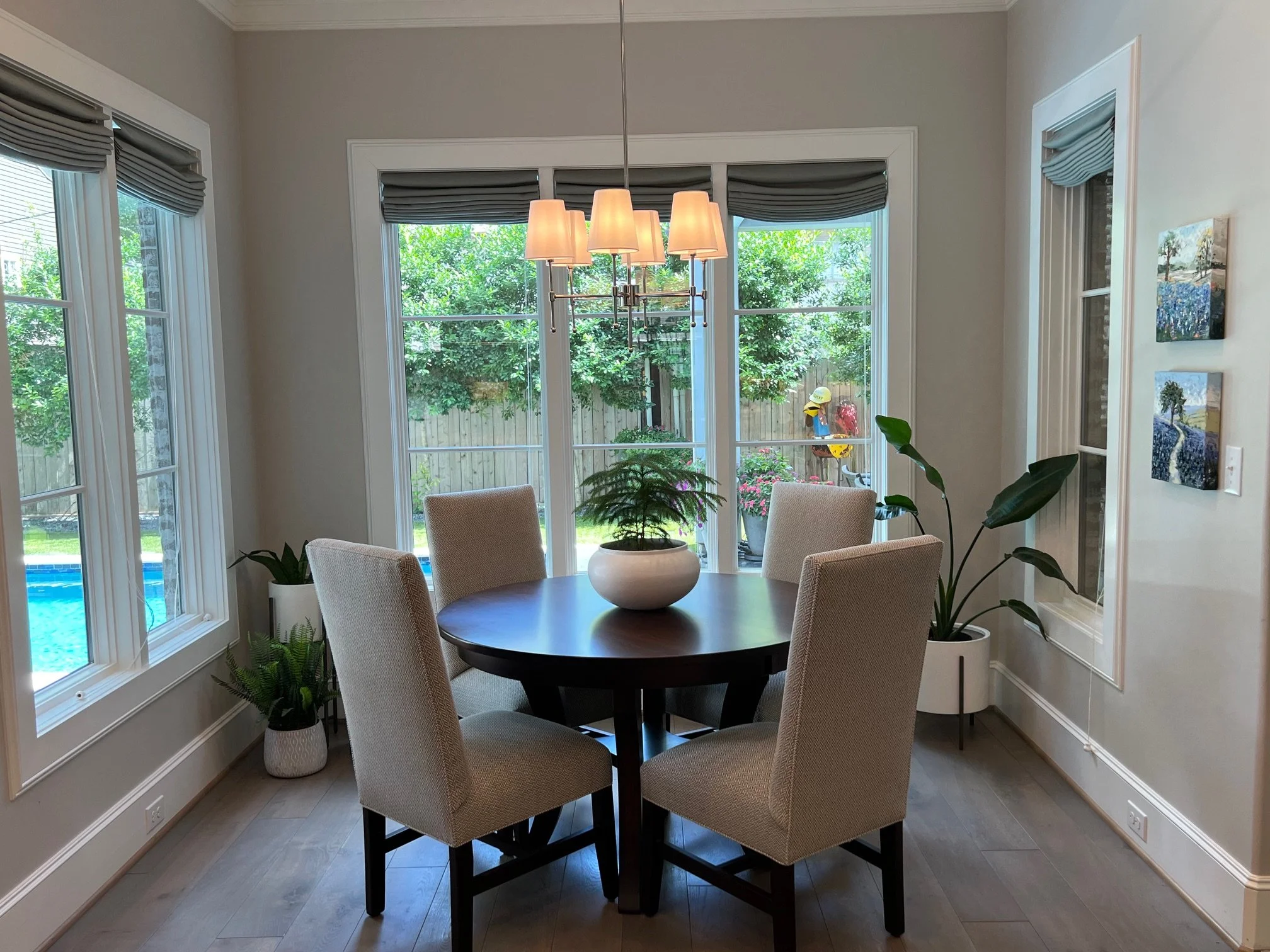 A round dining table with six upholstered beige chairs in a bright dining room with large windows, green trees outside, potted plants, and wall art.