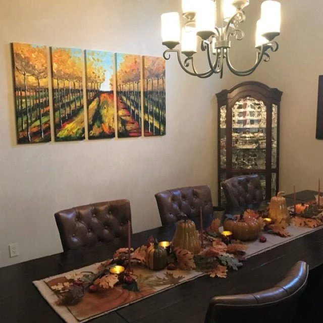 Dining room with a dark wooden table, six brown tufted chairs, autumn-themed table decor, a five-panel landscape painting of trees and a pathway, a glass-front cabinet, and a chandelier with five lights.