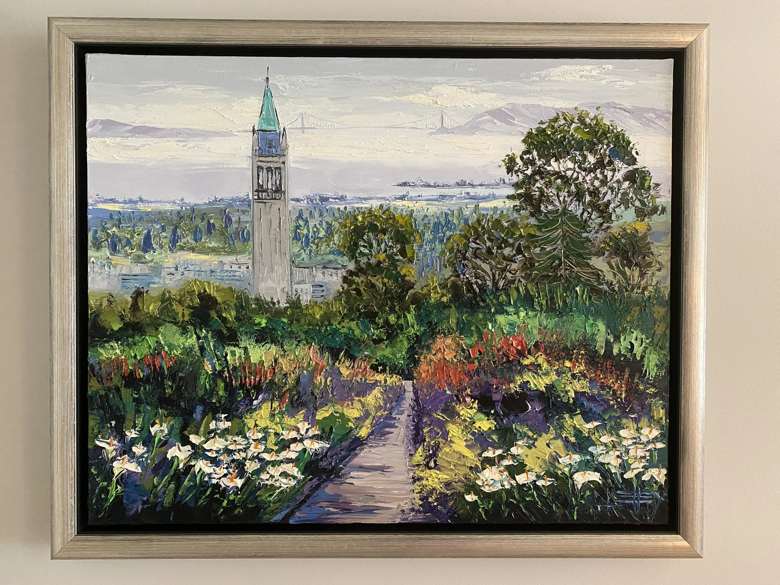 A framed impressionist painting of a church with a tall steeple, overlooking fields with a colorful flower garden in the foreground and mountains in the background.