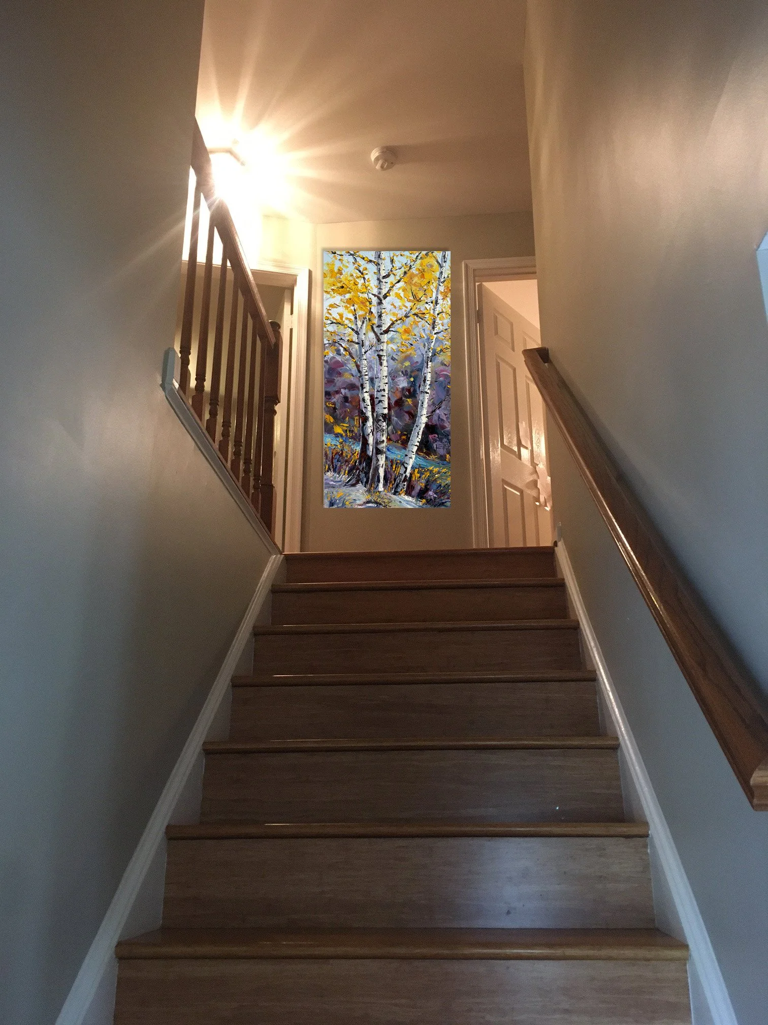 Interior view of a staircase with wooden steps, a wooden handrail, and a wall to the left. At the top of the stairs, there is a colorful painting of a forest scene featuring white birch trees with yellow leaves, set in a bright space with open doors.