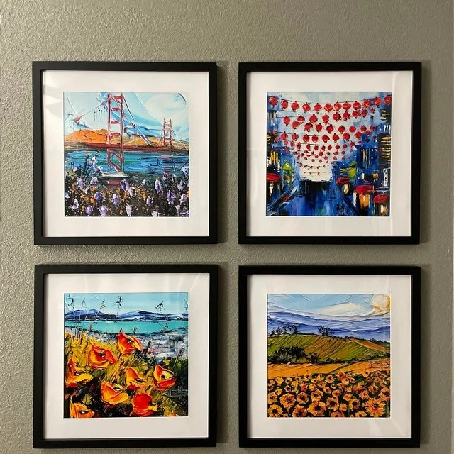 Four framed watercolor paintings on a gray wall, depicting the Golden Gate Bridge, a street with red lanterns, a field of orange poppies, and a landscape with rolling hills and flowers.