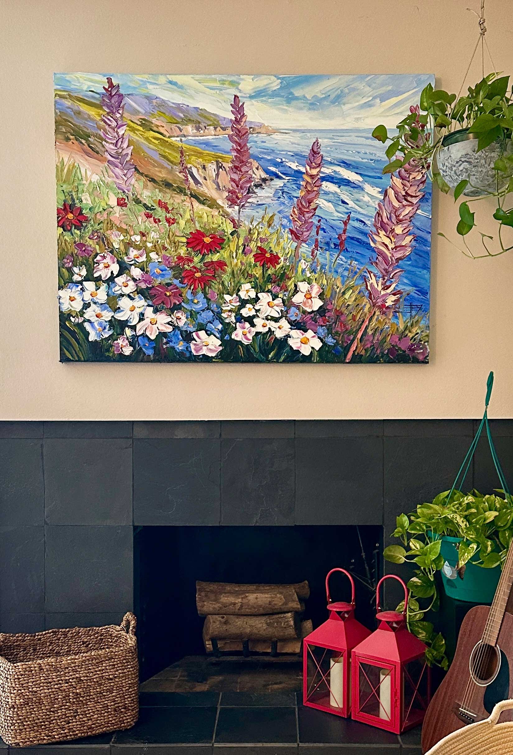 Painting of coastal landscape with cliffs, flowers, and ocean waves hanging above a fireplace, decorated with potted plants and red lanterns, in a cozy indoor space.
