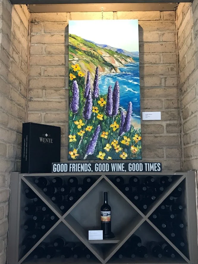 A colorful painting of a coastal landscape with purple and yellow flowers in the foreground, cliffs and the ocean in the background, hung on a brick wall in an art gallery. Below the painting is a sign that reads 'GOOD FRIENDS, GOOD WINE, GOOD TIMES'