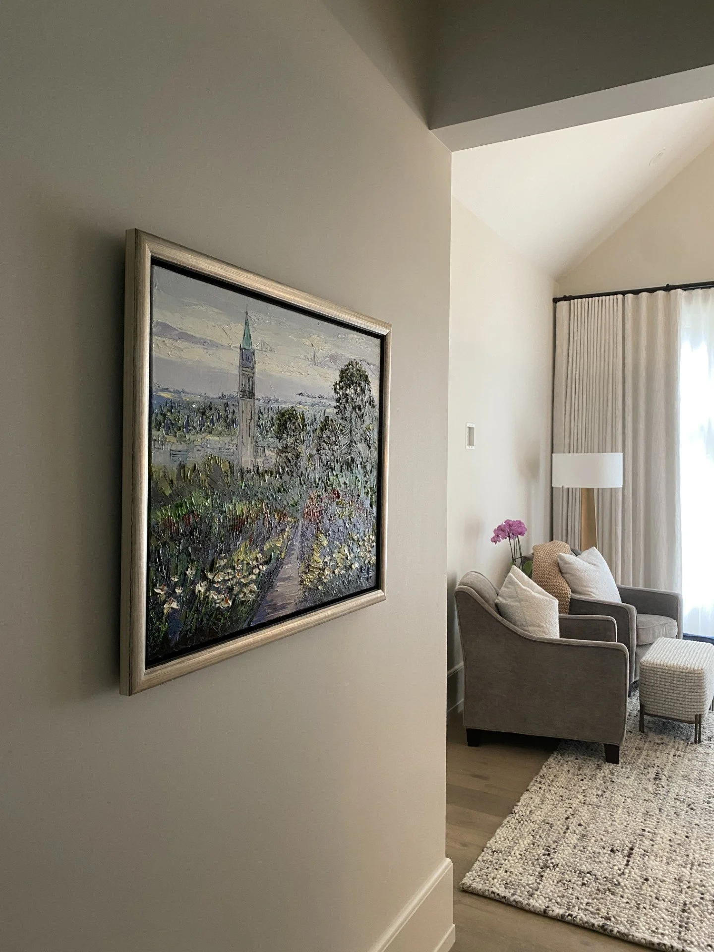 Living room with beige chairs, a floor lamp, a floral arrangement, window with curtains, and a framed painting on the wall.