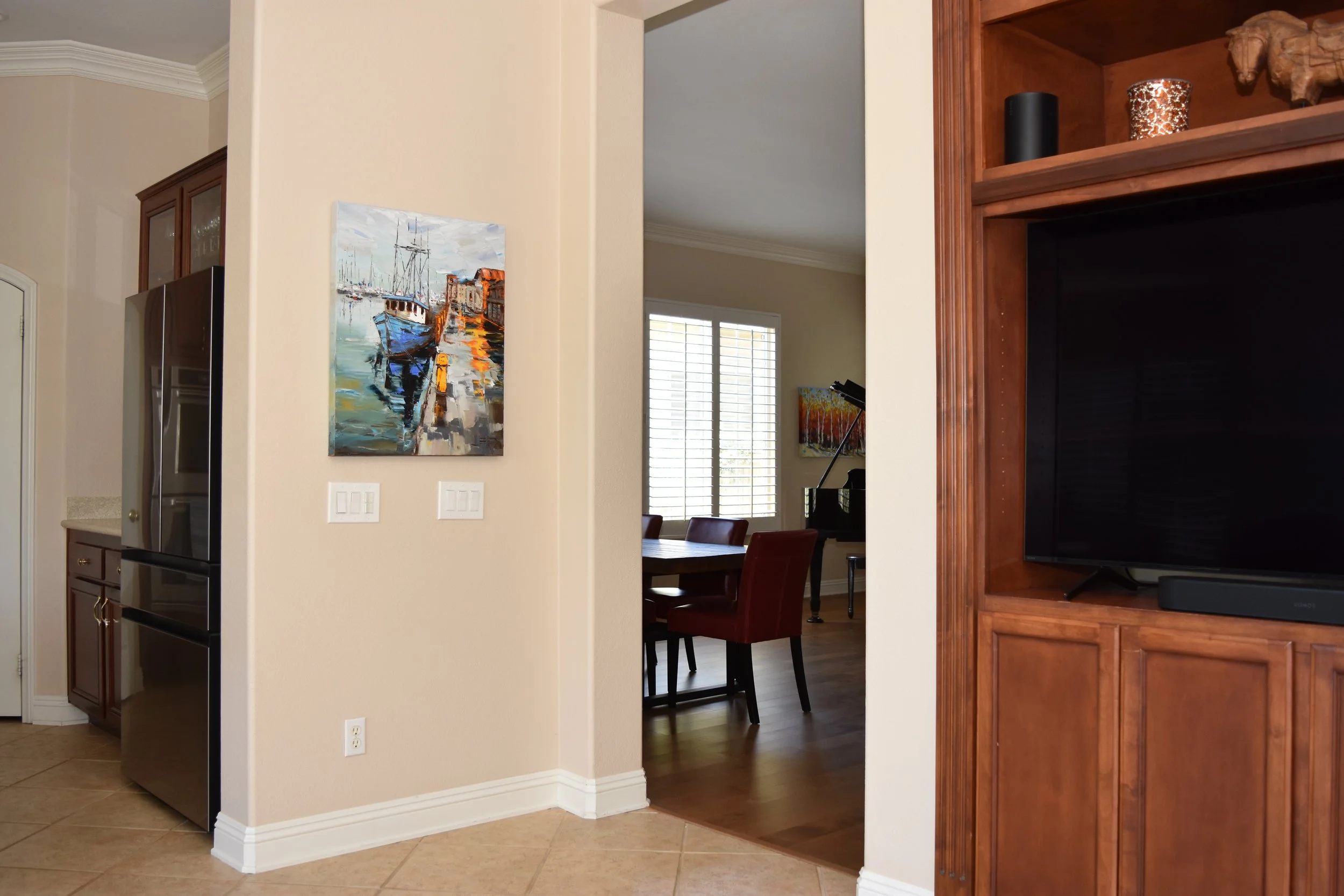 Interior view of a home showing a beige wall with a colorful boat painting, a doorway to a dining area with a wooden table and chairs, a window with shutters, and a built-in wooden entertainment center with a TV. A corner of the kitchen with a refrig