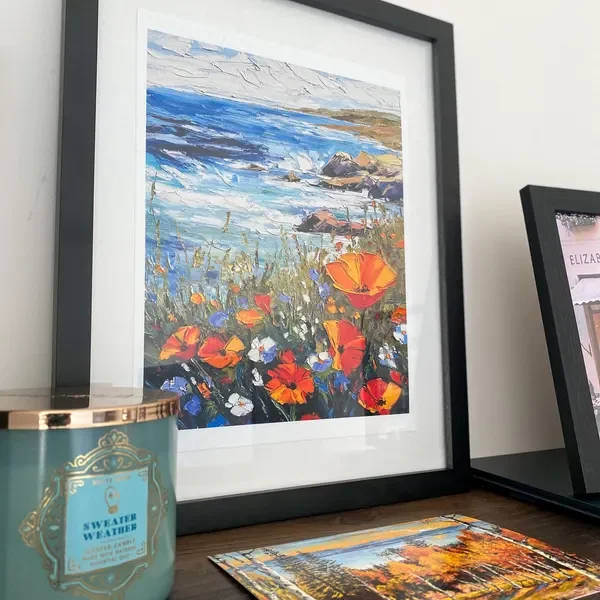 Framed landscape painting of a seascape with wildflowers in the foreground, placed on a table beside a candle and another framed picture.