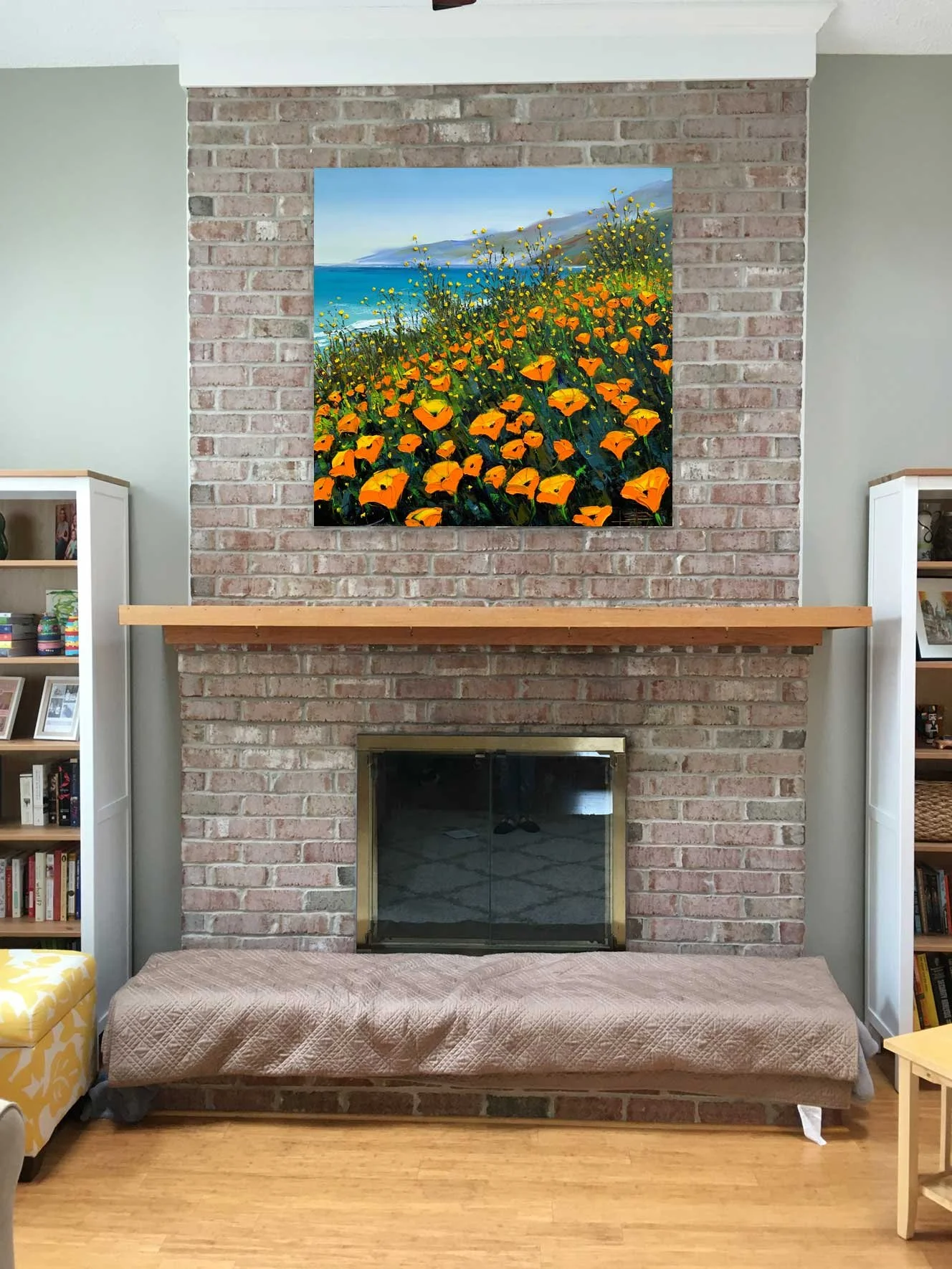 A living room with a brick fireplace, above which hangs a landscape painting of flowers in the foreground and mountains near the ocean in the background. There is a beige quilted couch in front of the fireplace, and symmetrical white bookshelves on e