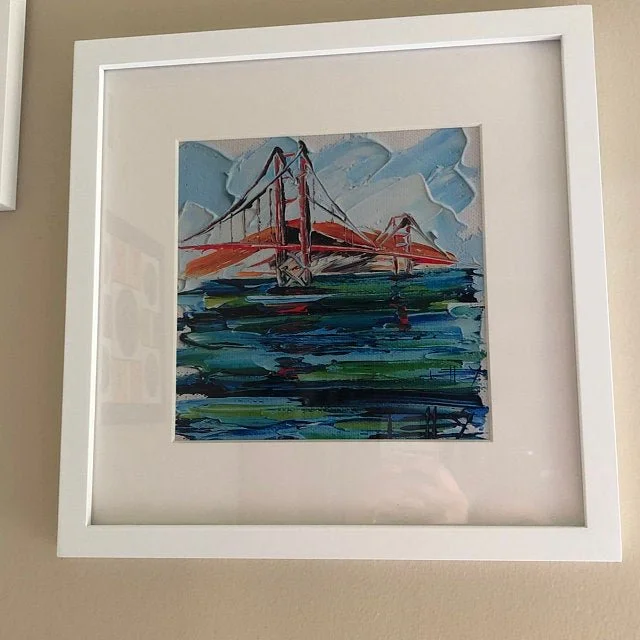 An abstract painting of the Golden Gate Bridge in San Francisco with a sky and water scene in shades of blue, green, red, and white, framed in a white square frame.