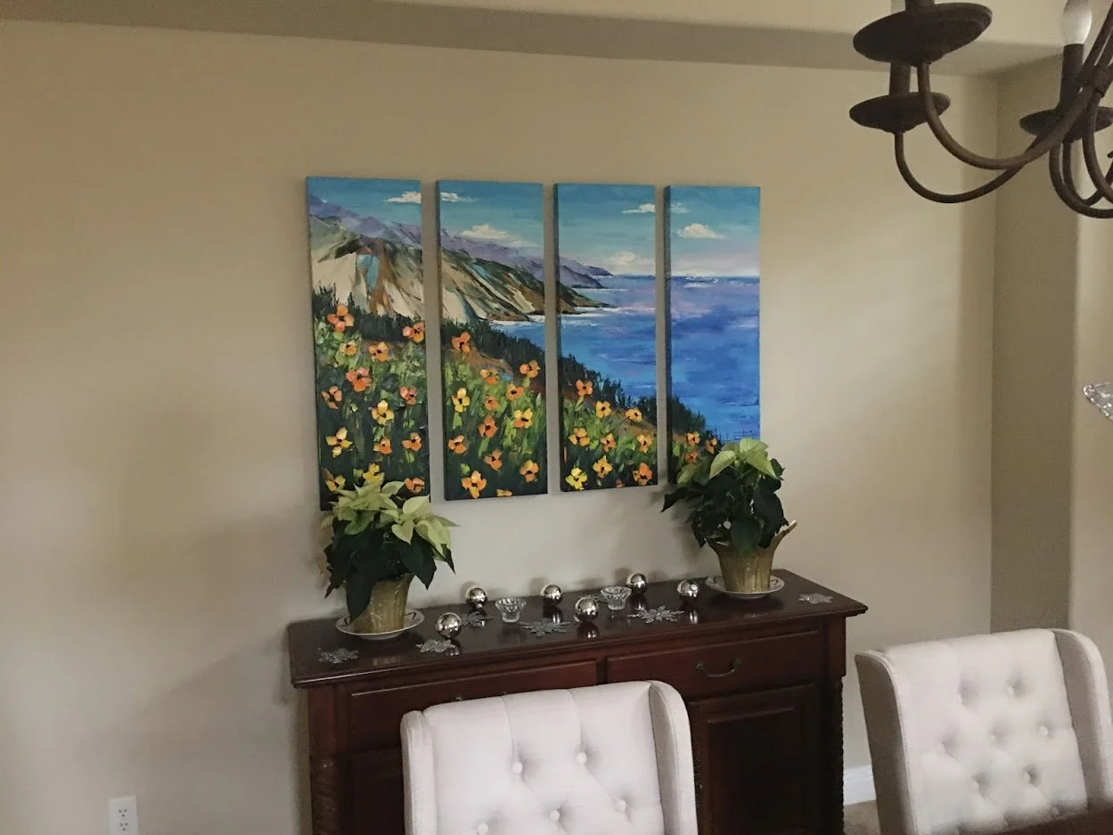 A multi-panel landscape painting of a coastal scene with mountains, ocean, and colorful flowers hangs above a wooden sideboard with decorative items and potted plants, in a room with beige walls and white upholstered chairs.