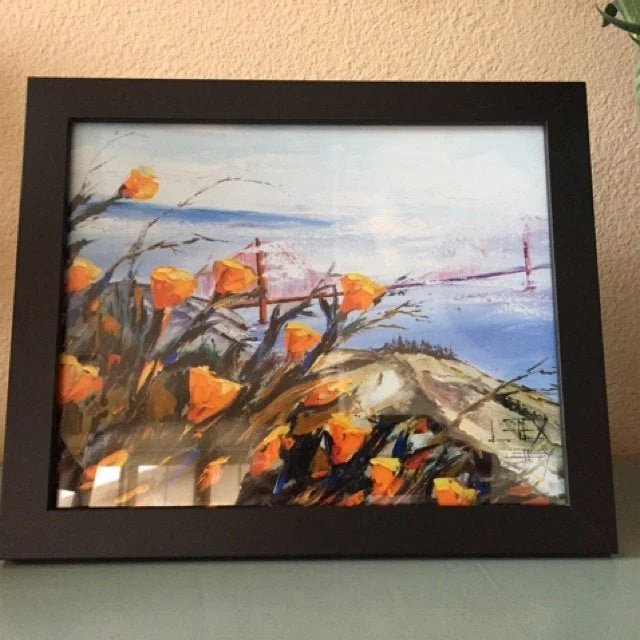 A framed painting of orange flowers and a landscape with a body of water and hills in the background.