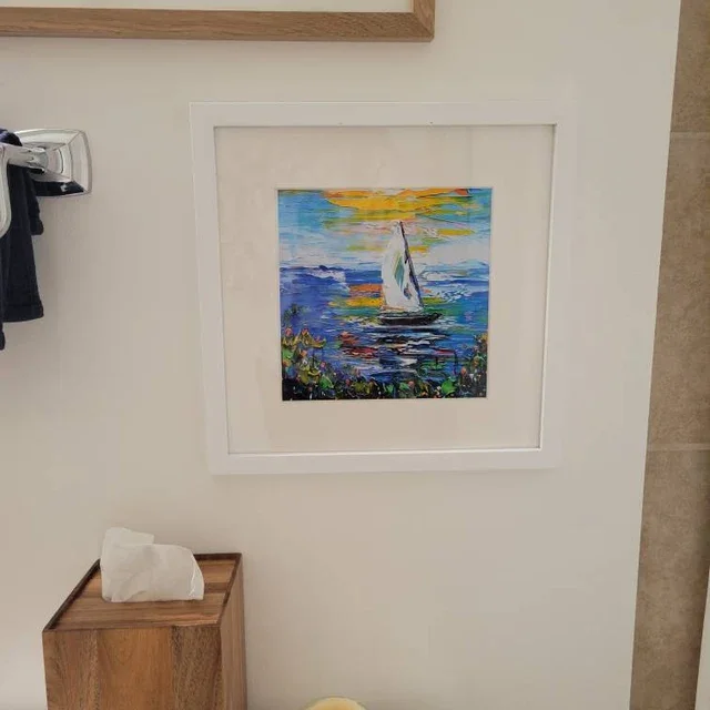 Framed painting of a sailboat on water with colorful sunset sky, hung on a wall next to a tissue box on a wooden table.