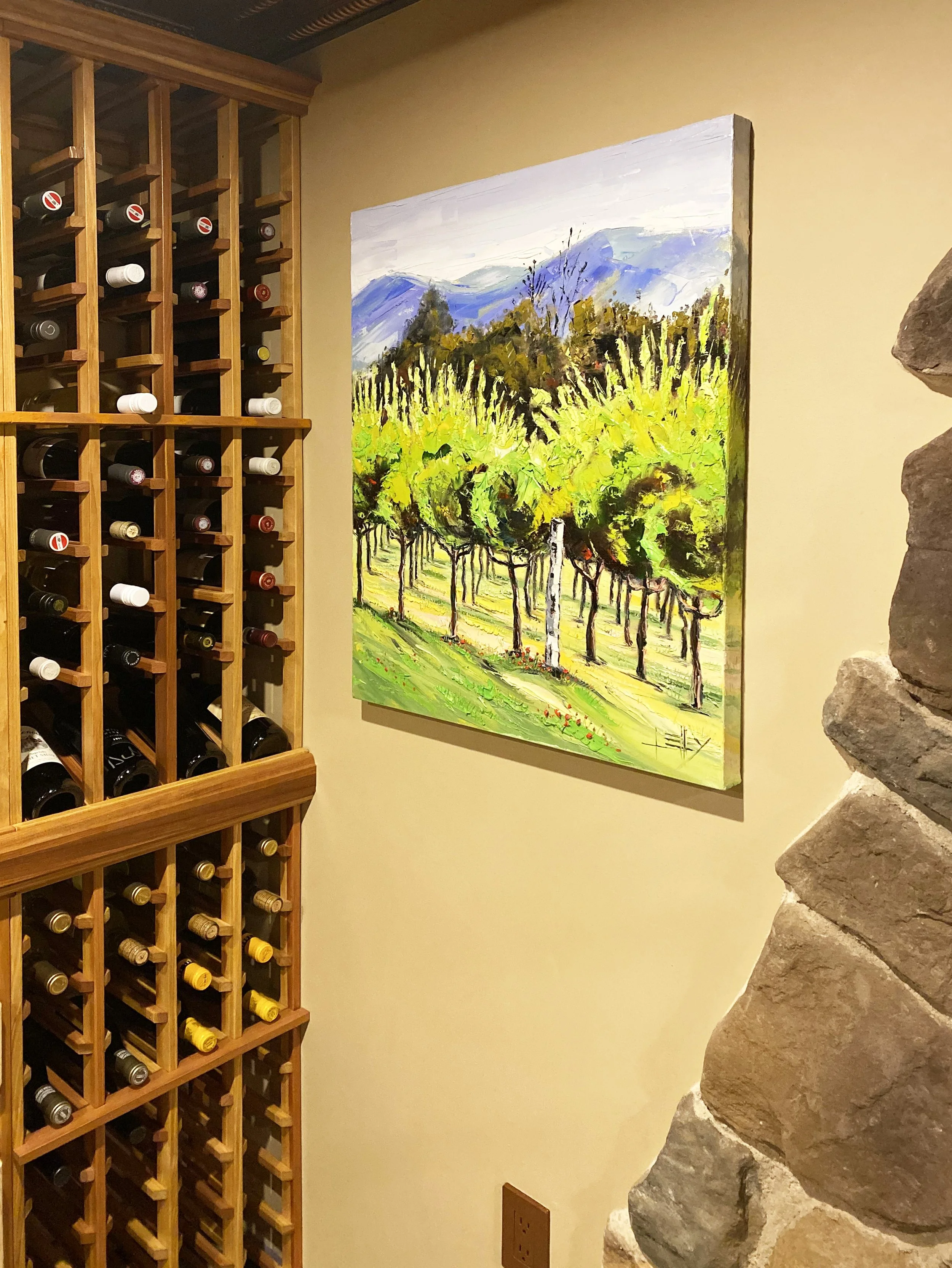 A wine rack filled with bottles on a wall next to a colorful landscape painting of mountains and trees on a wall.