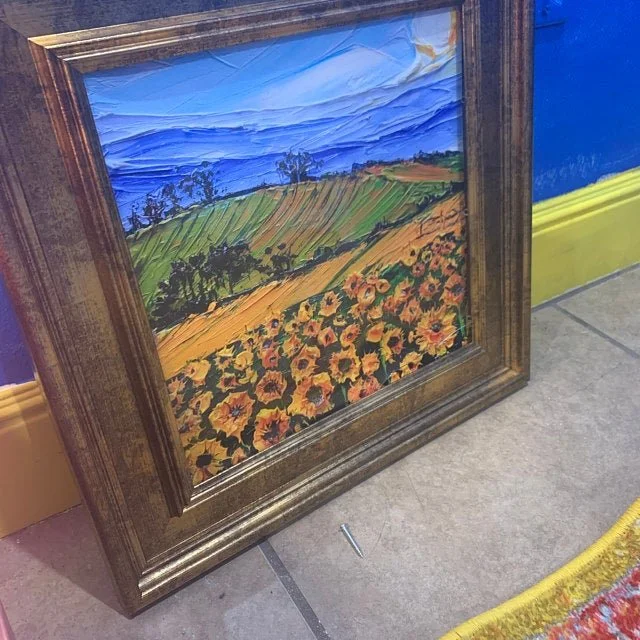 Framed painting of a landscape with green hills, a field of sunflowers, and a blue sky with clouds.
