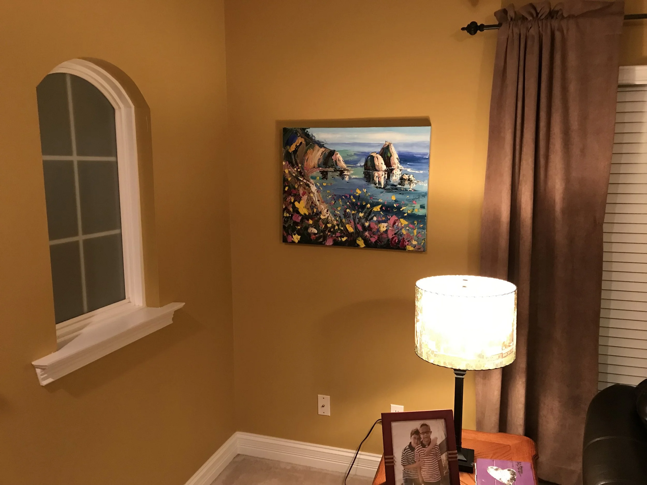 Living room with yellow walls, a colorful mountain lake landscape painting, brown curtains, a floor lamp with a beige shade, a wooden console table, framed photographs, a laptop, and a window with white blinds.