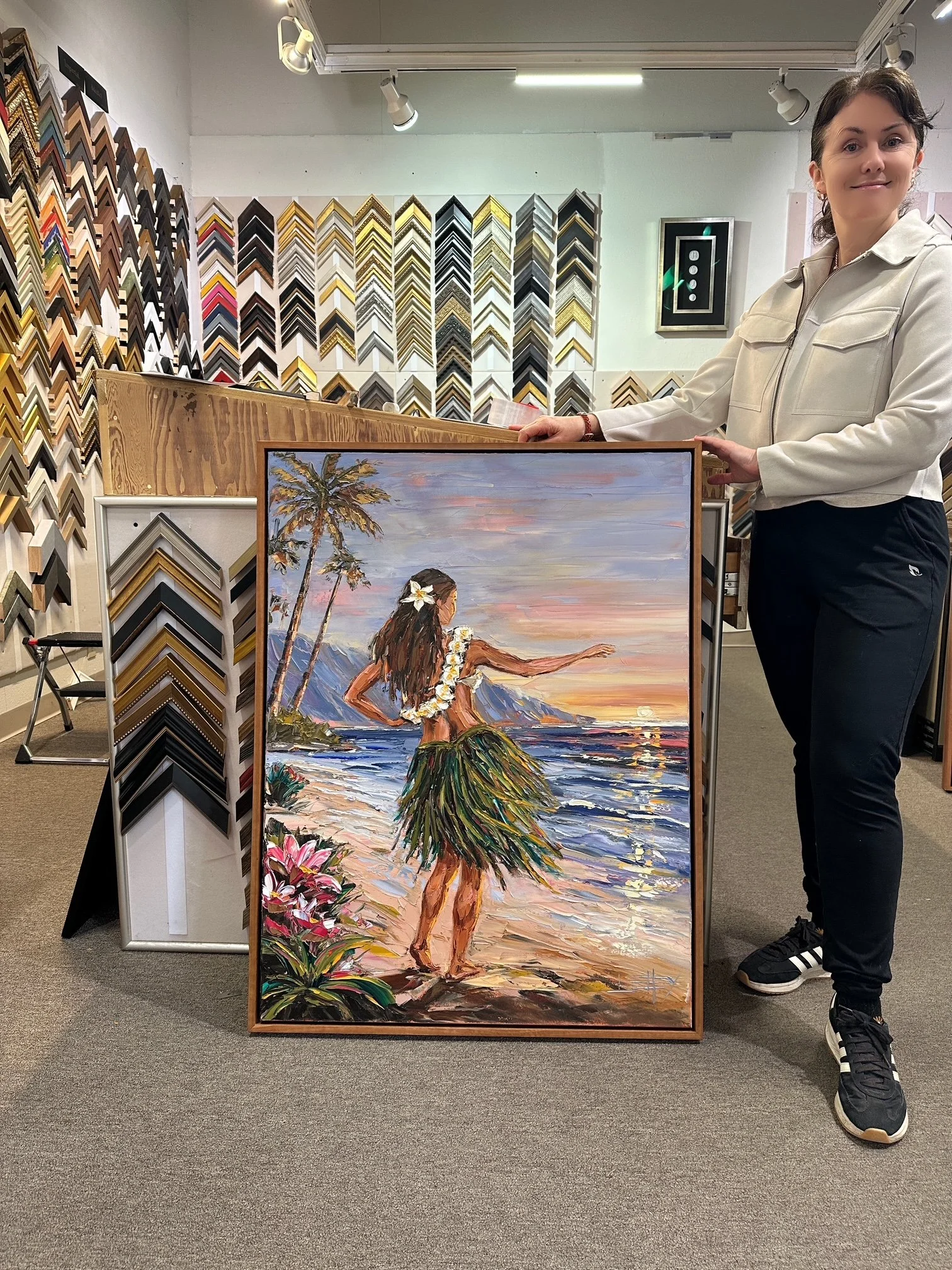 A woman in a light-colored jacket and black pants holding a painting of a hula dancer on a beach at sunset, with palm trees and flowers, inside an art supply store with framed picture samples on the wall.