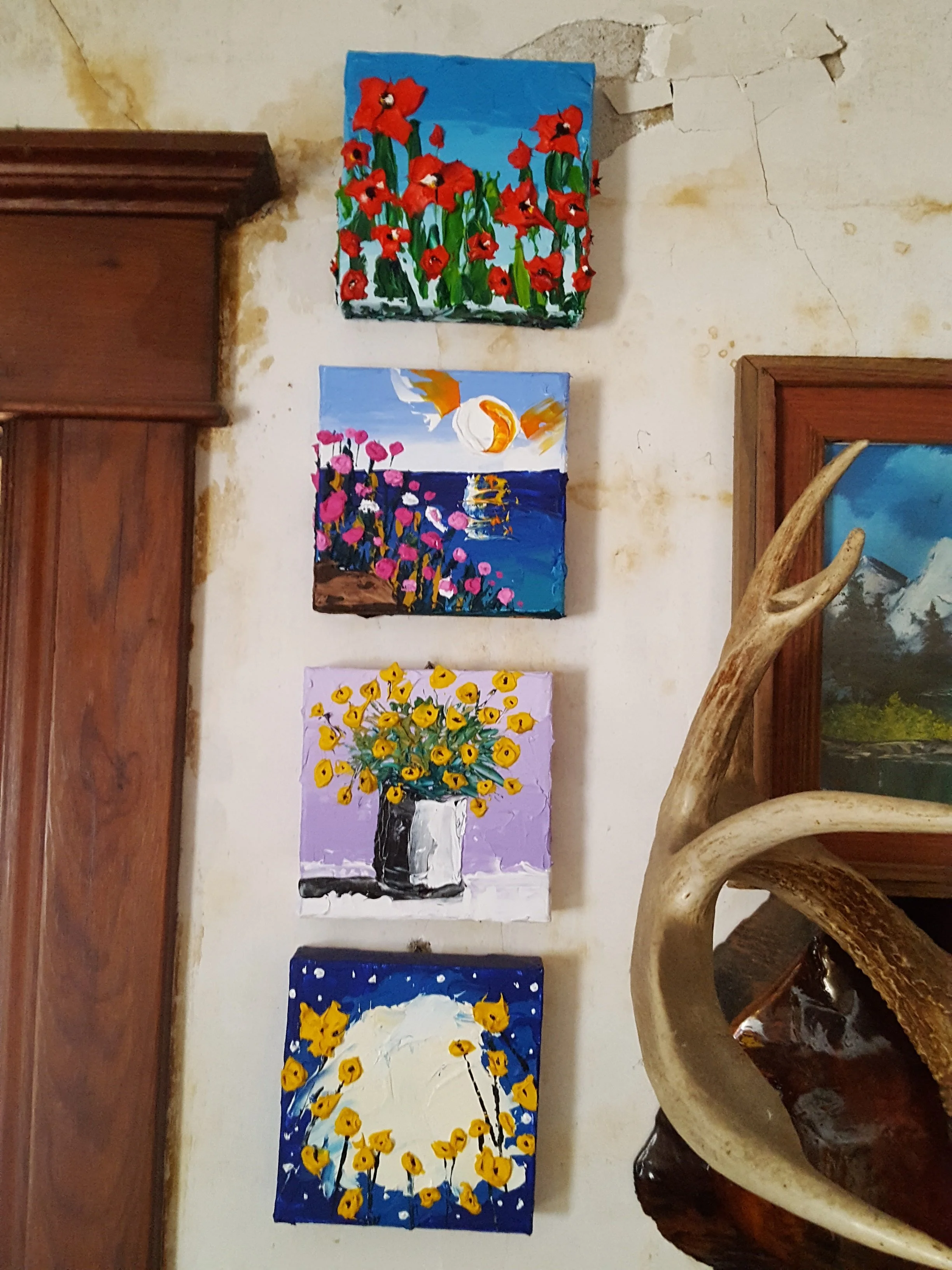 Four colorful paintings of flowers on a wall, with a wooden carved furniture piece and decorative animal horn in the foreground.