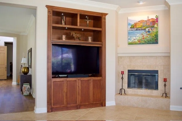 Living room with a large wooden entertainment center, a flat-screen TV, a fireplace with red candle holders, and a colorful landscape painting above the fireplace.