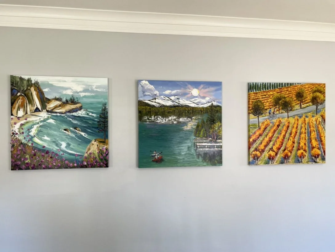 Three landscape paintings hanging on a white wall in a gallery or room. The paintings depict coastal, mountain, and vineyard scenes.