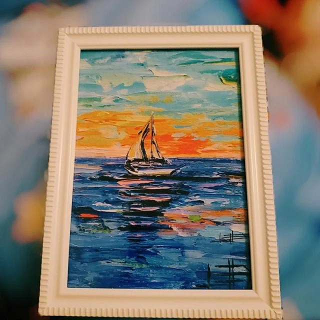 A framed oil painting of a sailboat on the water at sunset, with vibrant orange, yellow, and blue sky, and reflections on the waves.