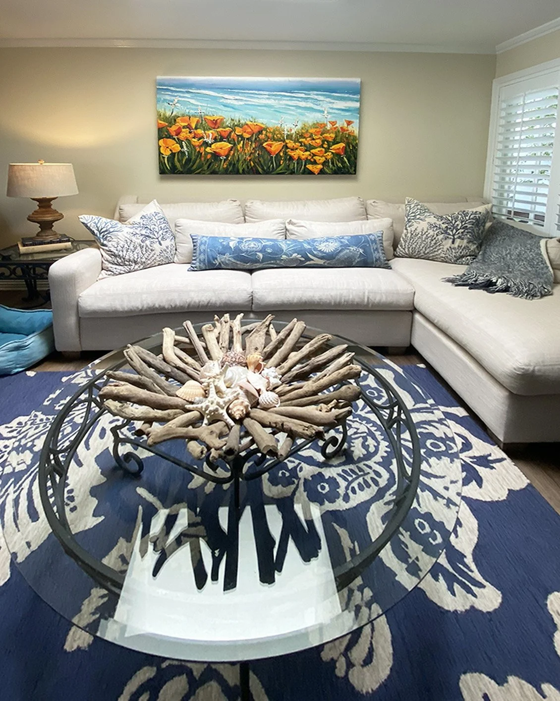 A living room with a cream-colored sectional sofa, decorative pillows, a blue and white patterned rug, a glass-topped coffee table with driftwood and seashell decor, a wall painting of a flower field, a table lamp, and a window with blinds.