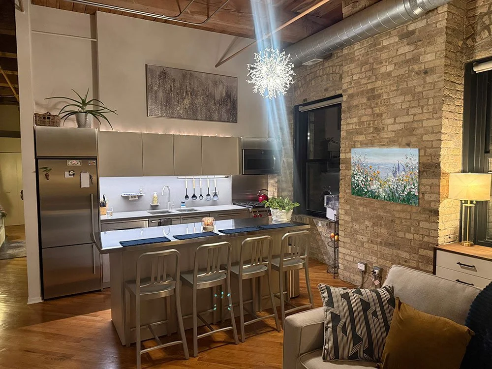 A modern kitchen with a brick wall, stainless steel refrigerator, light grey cabinets, a white countertop with a sink, and a row of four bar stools at a kitchen island. There is a snowflake-shaped light fixture hanging from the ceiling, a window with black framing, a painting of flowers on the brick wall, and a beige sofa with black and gold pillows nearby.