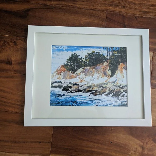 Framed painting of a coastal landscape with cliffs, trees, and ocean waves.
