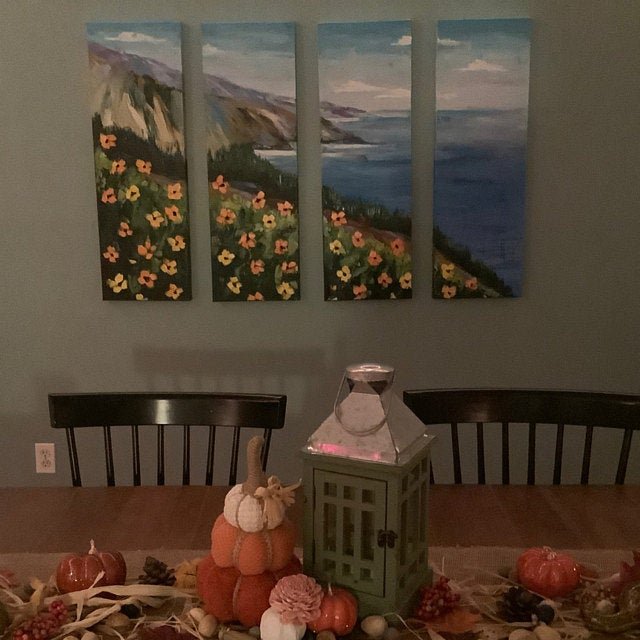 A dining room with a wooden table decorated for fall, featuring pumpkins, pinecones, and flowers. A gray lantern and glass terrarium are on the table. Four-panel landscape painting of a coastal scene with cliffs, flowers, and ocean hangs on a teal wa