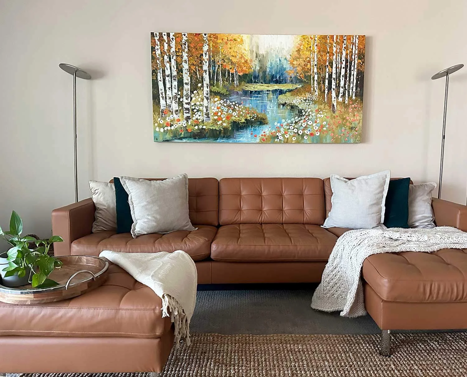 Living room with a tan leather sectional sofa with pillows, a knit throw, and a side table with a plant, above a colorful landscape painting of a river surrounded by trees.