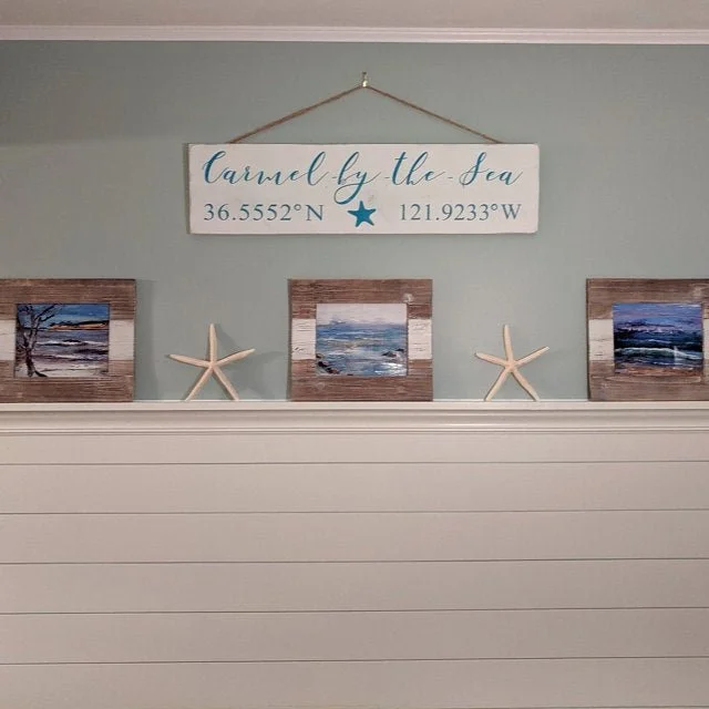 Beach-themed wall decor with a sign reading 'Caramel by the Sea' and displaying coordinates 36.5552°N, 121.9233°W, three framed seaside paintings, and four starfish on a wall.
