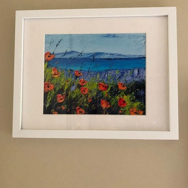 Framed landscape painting featuring red poppies in a field with water and mountains in the background.