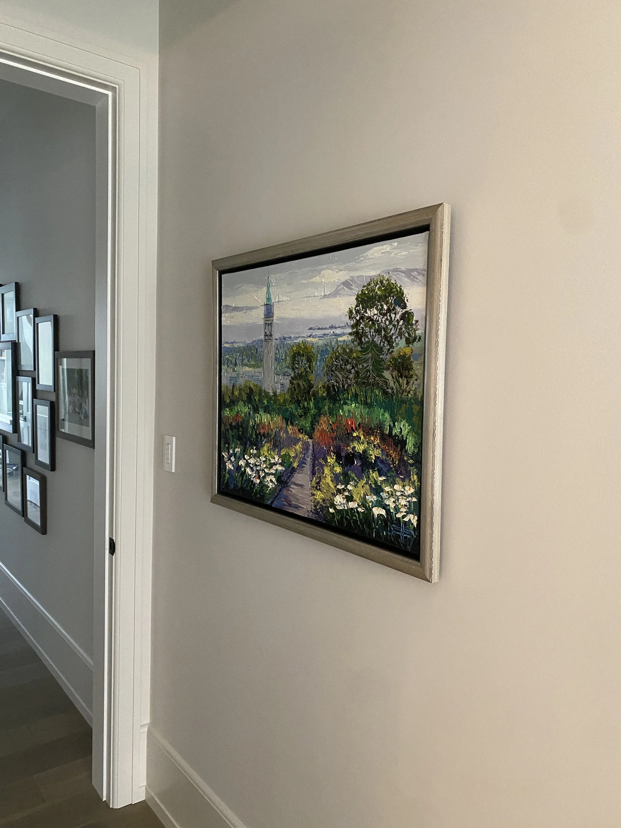 Interior view of a hallway with a painting of a scenic landscape featuring a church steeple, trees, colorful flowers, mountains, and sky, hung on a beige wall.