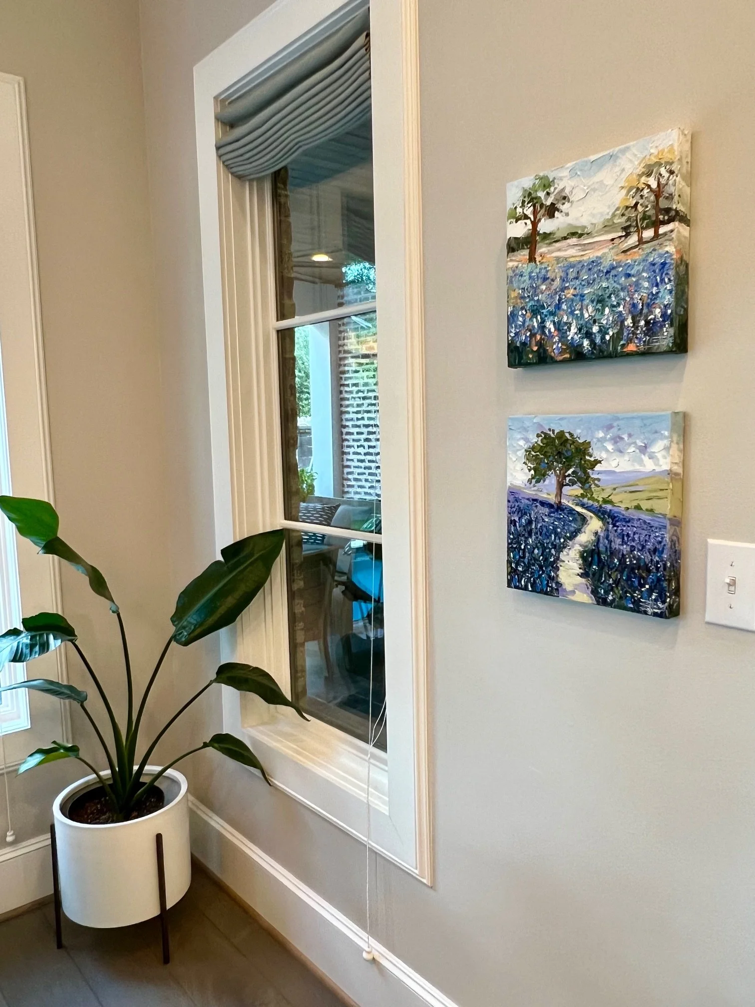 Interior view of a room with a plant in a white pot, a window with a white frame, and two landscape paintings on the wall. The window has a partially pulled blue and tan curtain, and outside the window, brick and greenery are visible.