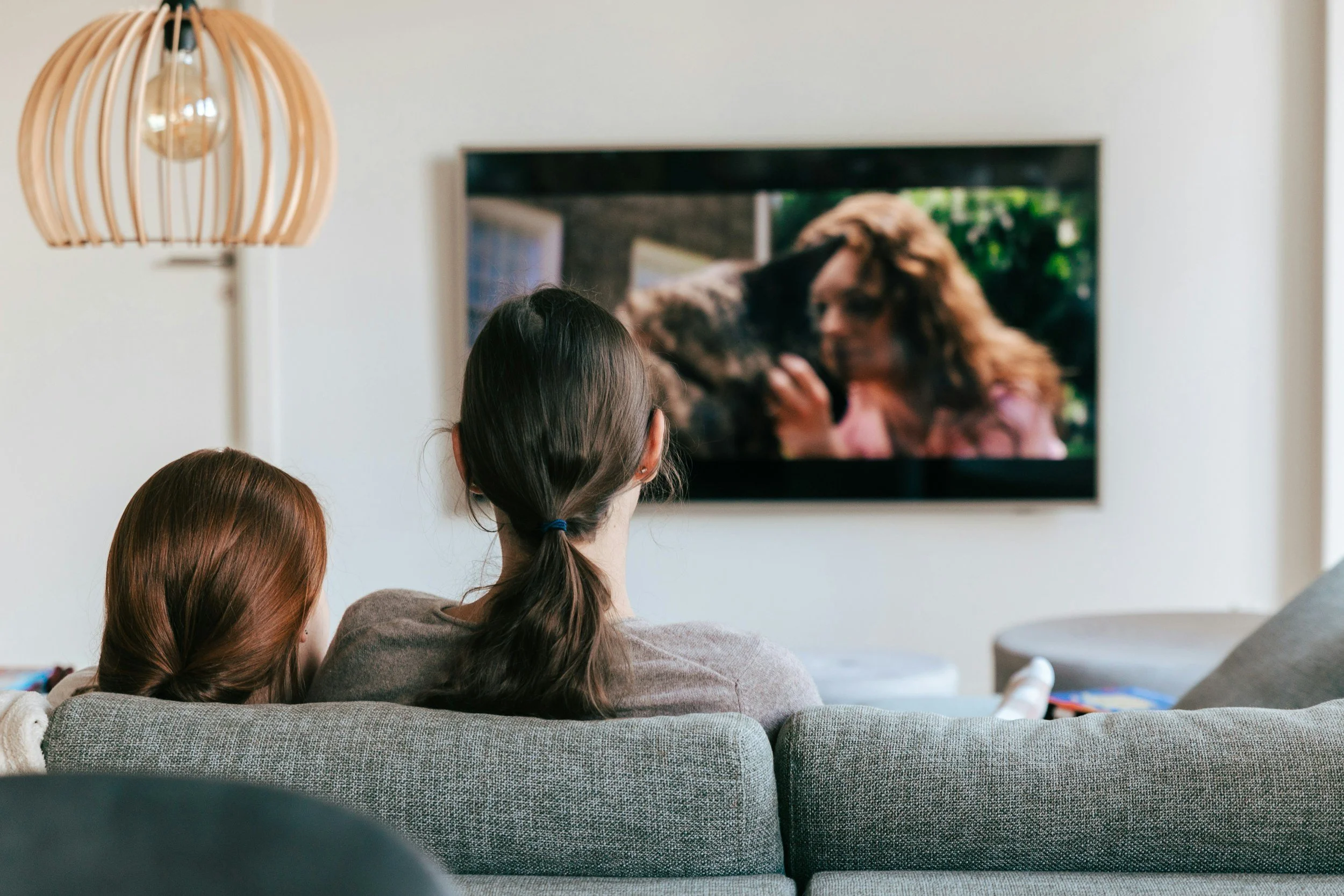 Two women with brown and reddish hair watching a movie or TV show on a flat screen in a cozy living room.