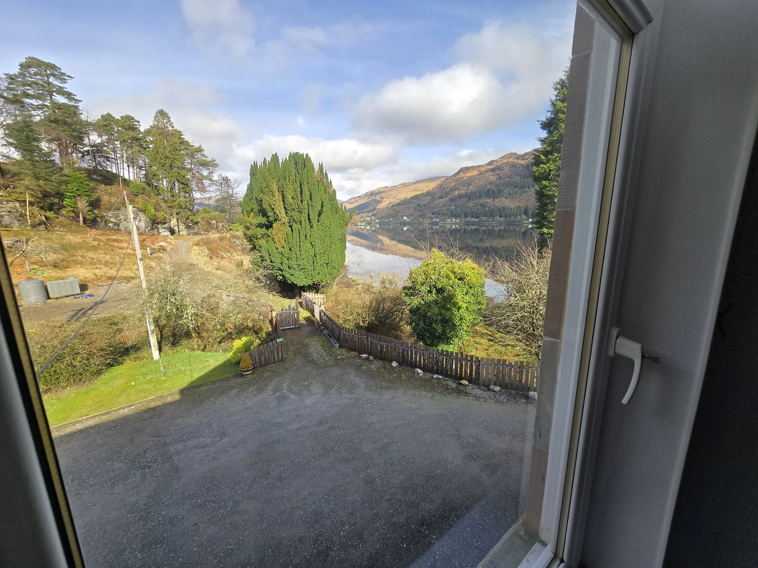 View from a window overlooking a rural landscape with a lake, hills, trees, a driveway, and a wooden fence.