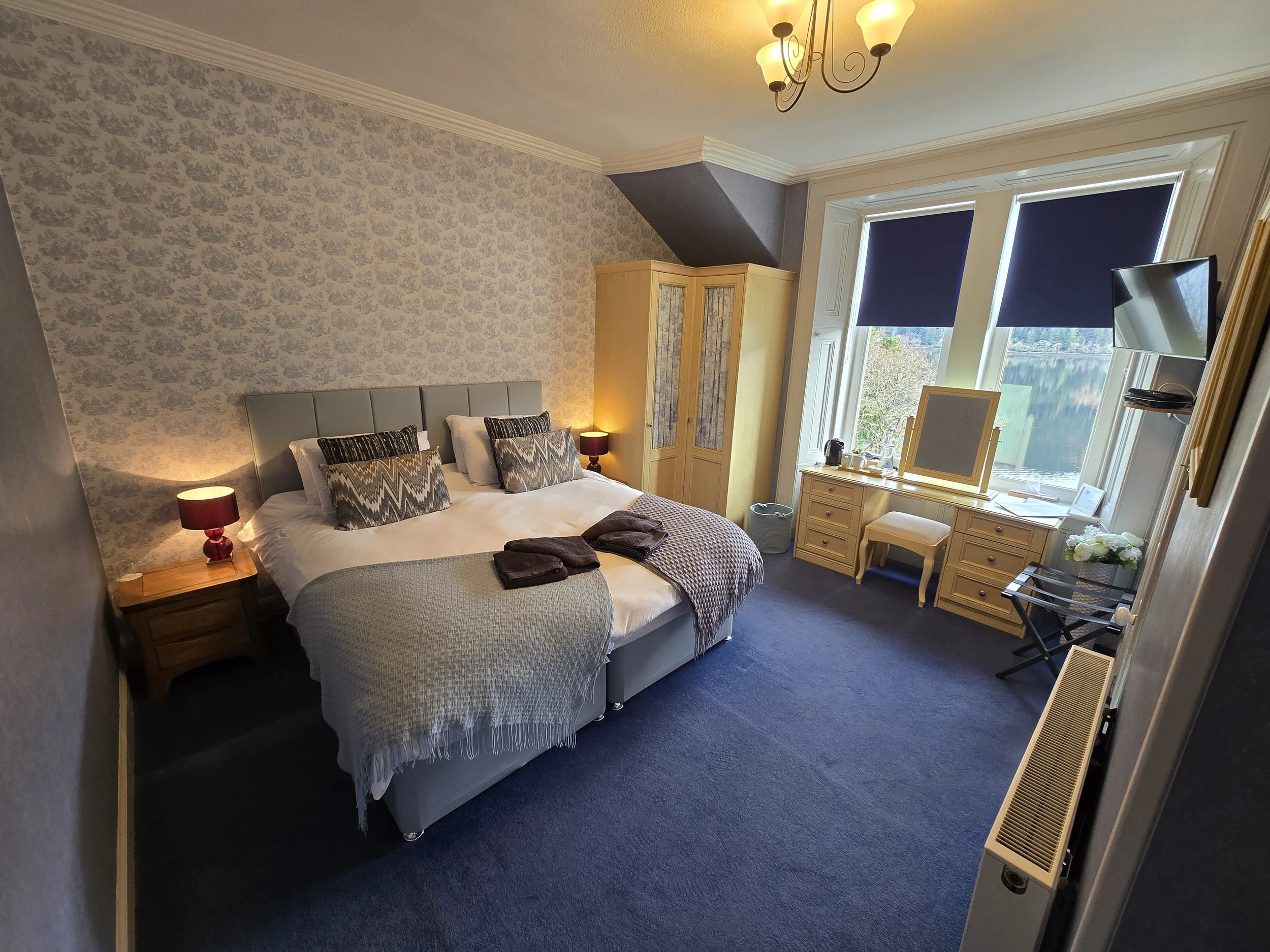 A cozy bedroom featuring a large bed with a padded headboard, pillows, and blankets, with bedside lamps, a wooden wardrobe, a desk by the window, a small stool, a wall-mounted TV, and large windows with blue blinds overlooking a scenic view of water and trees.