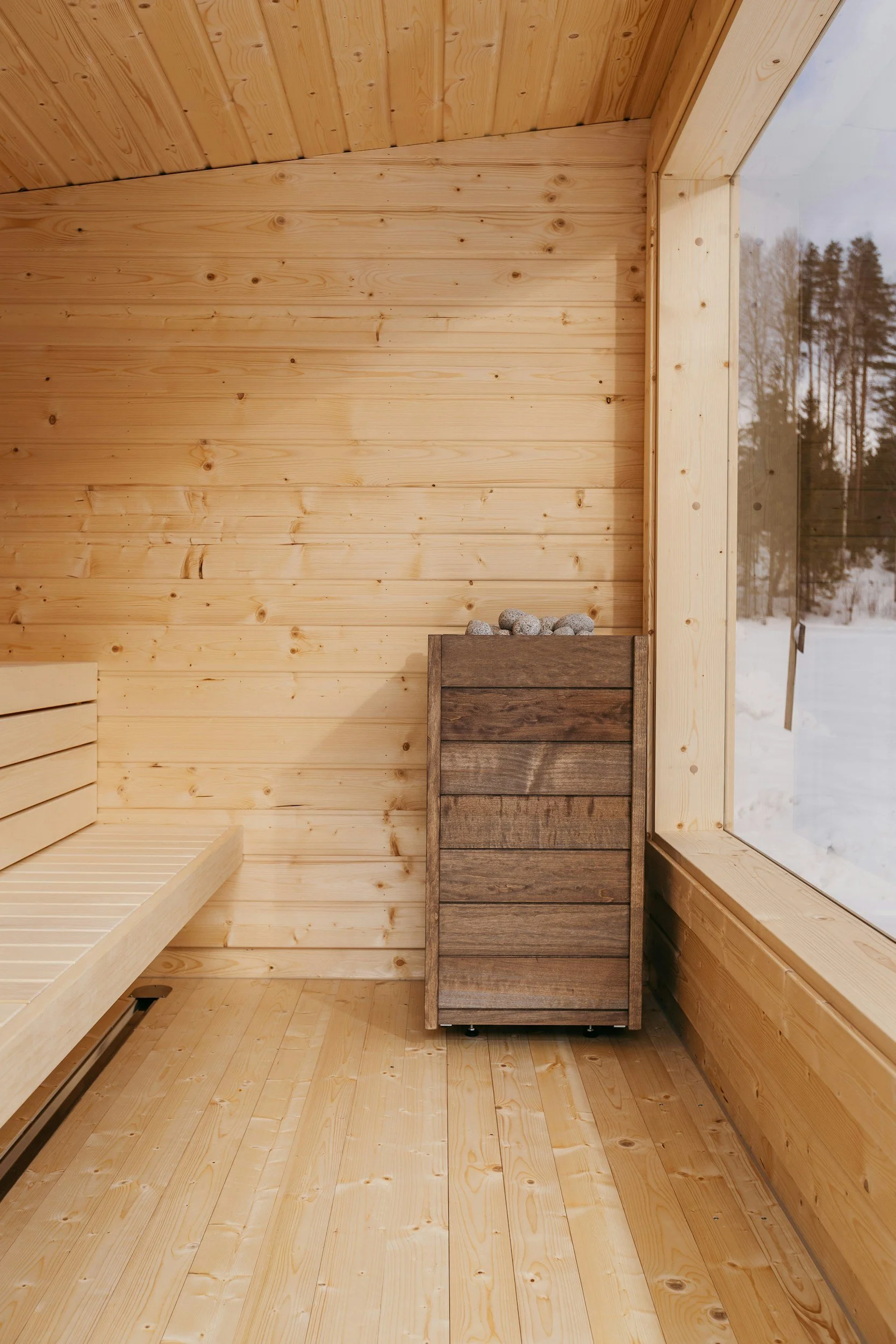 Interior of a wooden sauna with a wooden bench on the left and a small wooden heater on the right topped with rocks, next to a large window showing snowy landscape outside.