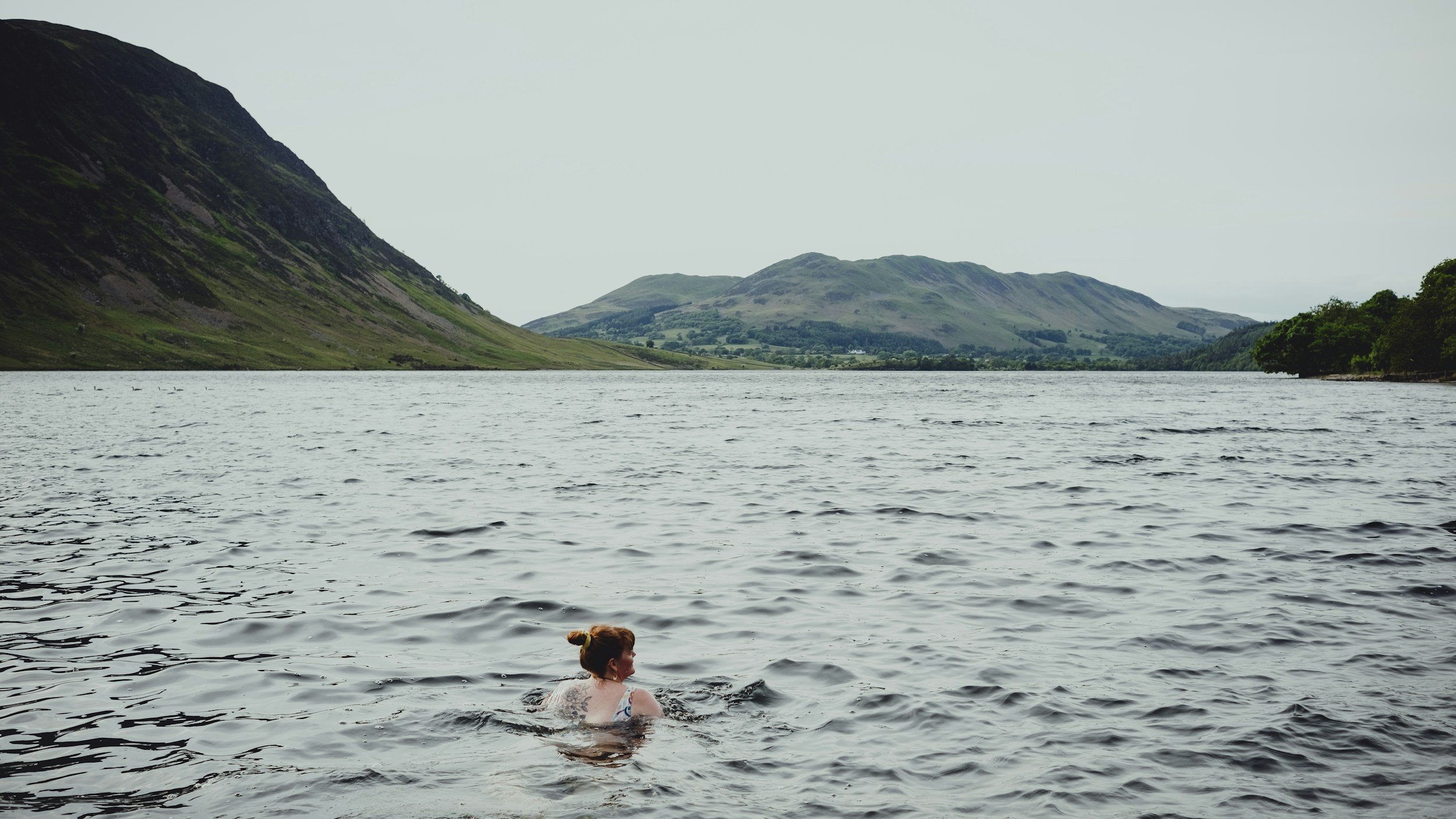 A woman swimming in a lake surrounded by green mountains with an overcast sky.