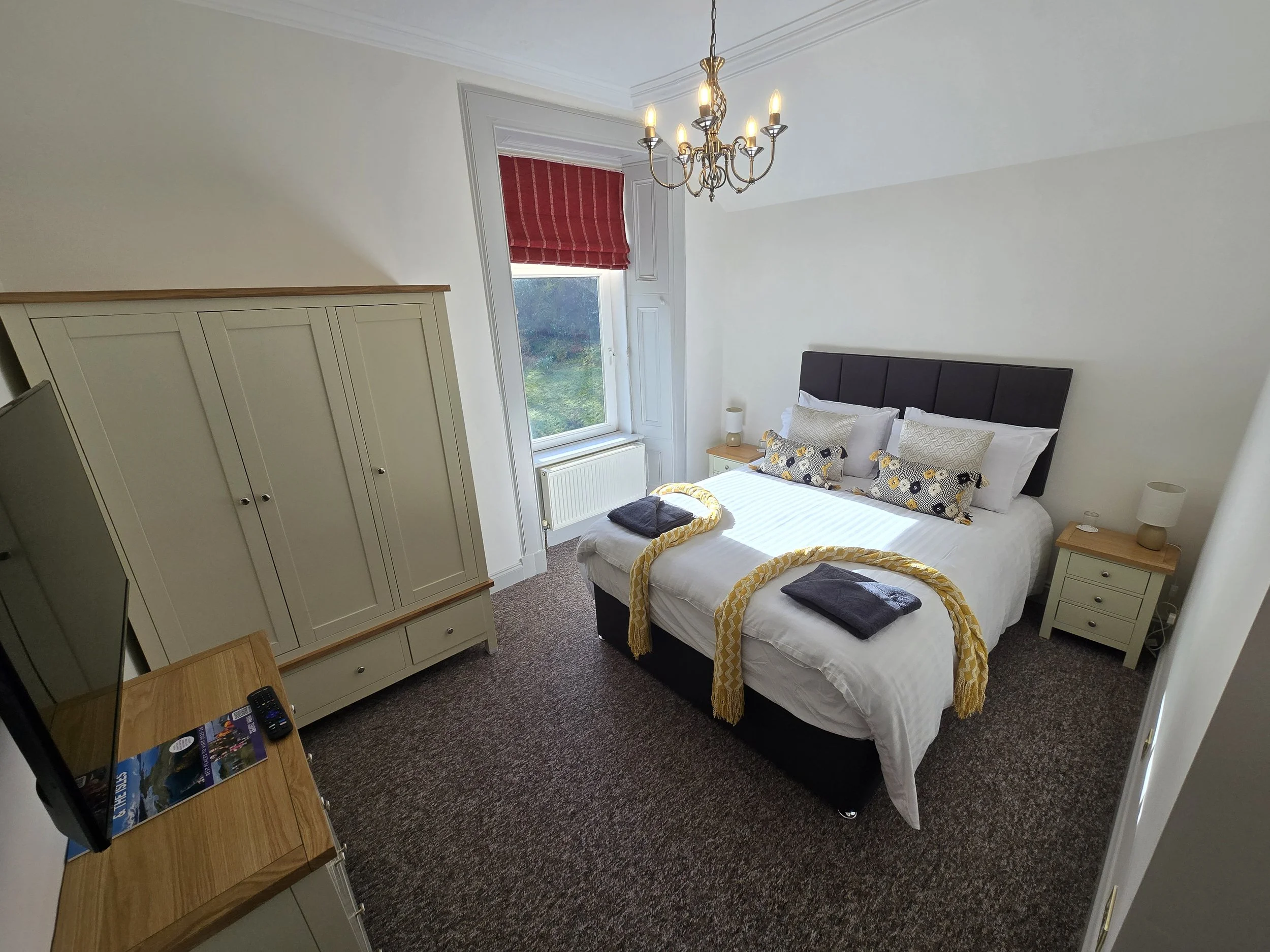 A bedroom with a bed, two nightstands, a wardrobe, a window with a red blind, a chandelier, and a TV on a wooden stand.