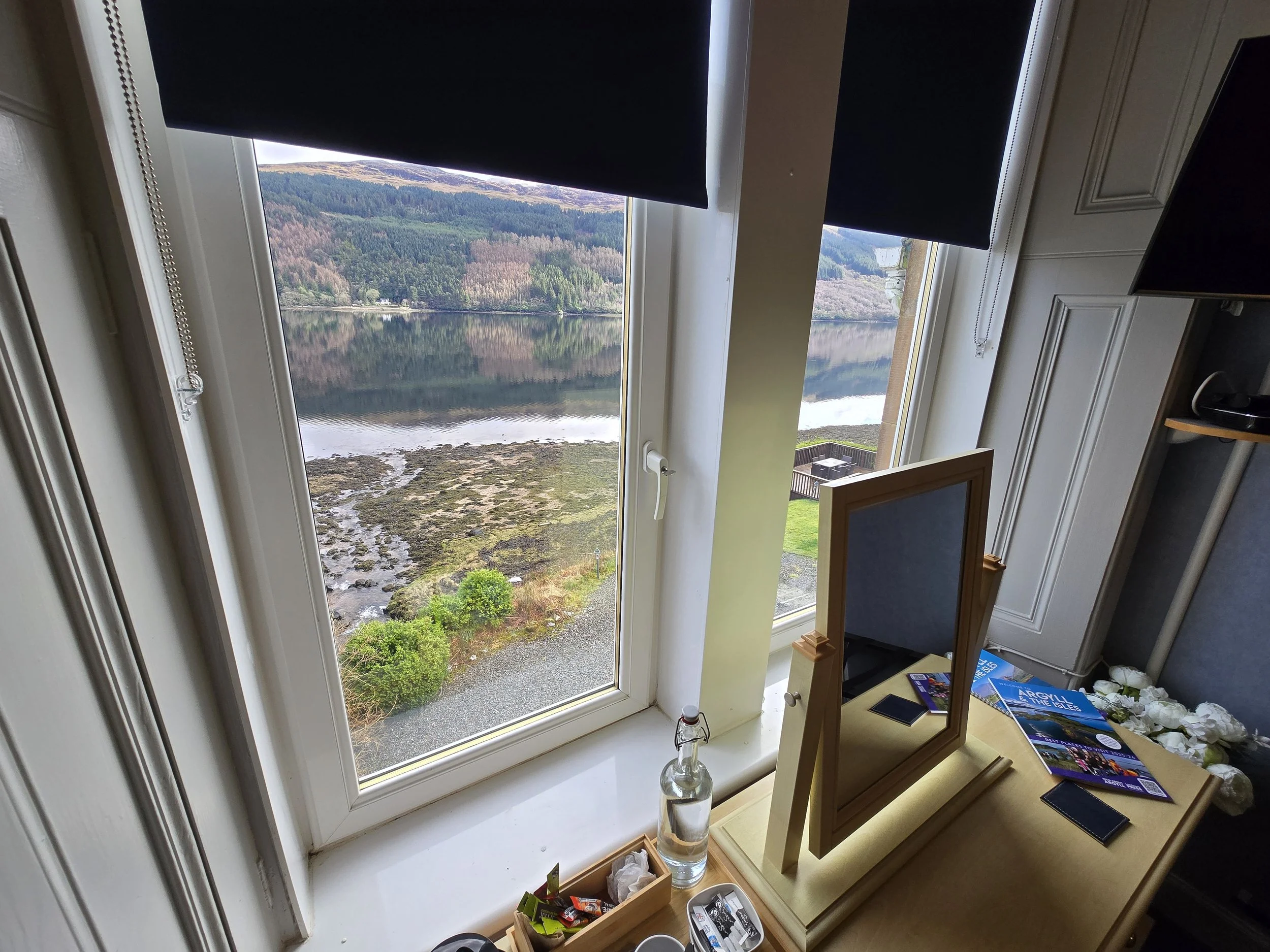 Room with a large window showing a scenic lake and hillside landscape, with a small makeup mirror, magazines, and bottles on a wooden dresser.