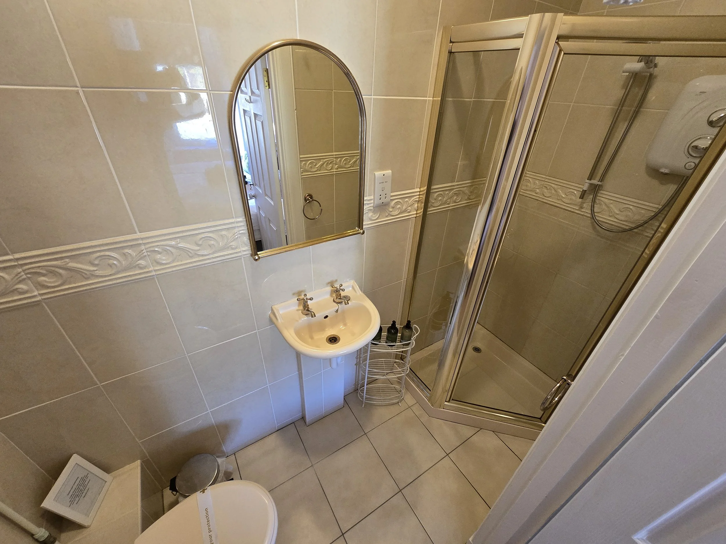 Small bathroom with beige tiled walls and floor, a white sink with brass faucets, an oval mirror above the sink, a shower with a glass door, a wall-mounted showerhead, and a toilet with a small trash can beside it.