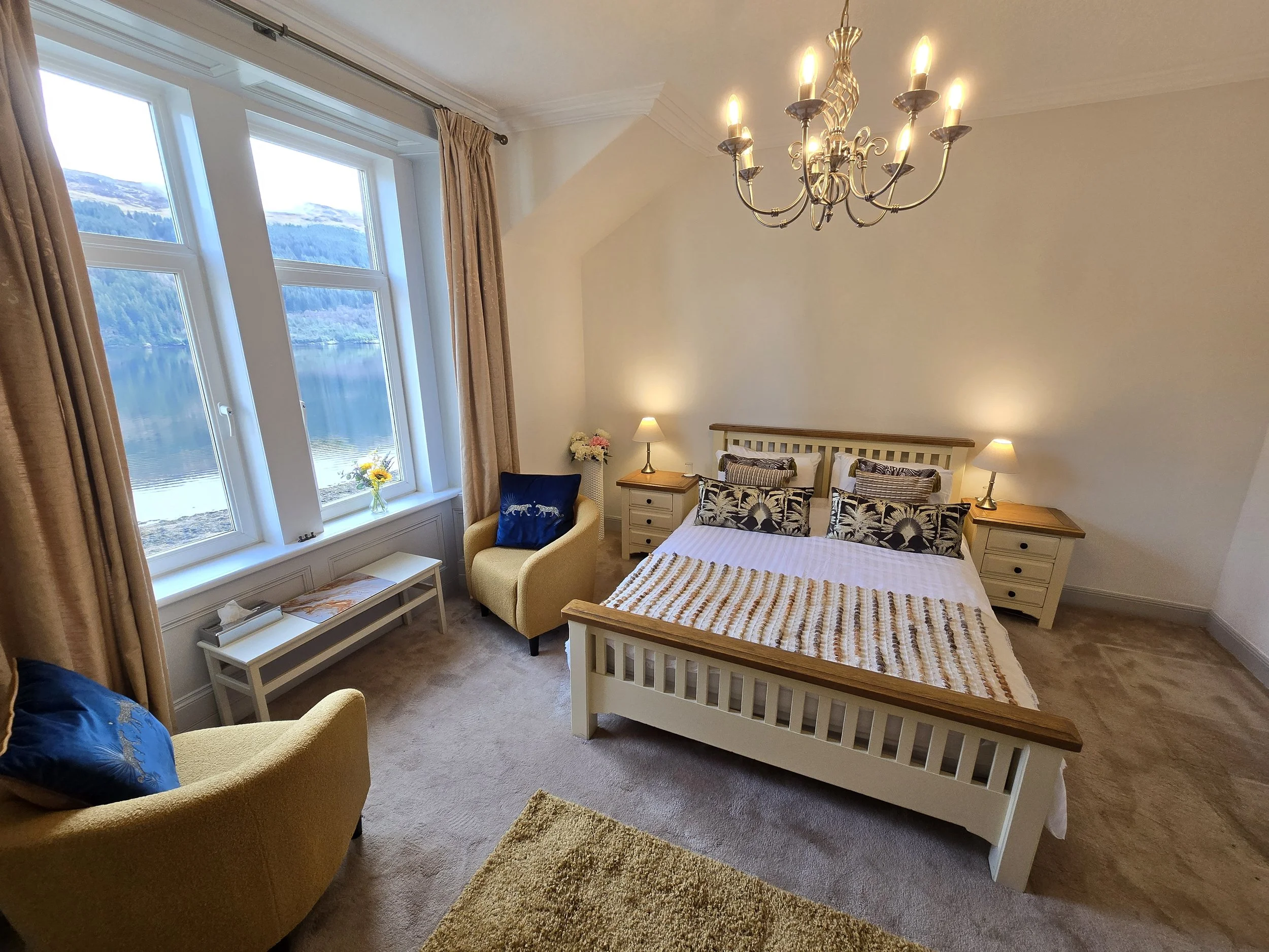 A cozy bedroom with a large window overlooking a lake and mountains, a white wooden bed with patterned pillows, two nightstands with lamps, a chandelier, a beige armchair with a blue pillow, and a small bench with flowers near the window.